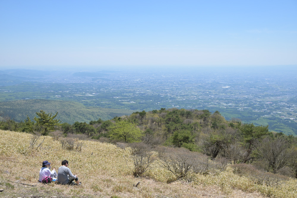 鍋割山の山頂から街並みを一望する登山夫婦と、晴天に広がる開放的な景色の風景