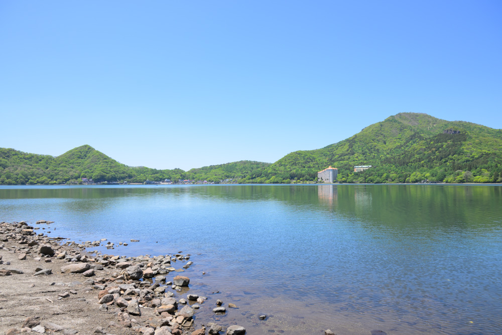 青空の下に広がる榛名湖畔の景色。澄んだ湖と新緑、爽やかな空が美しく、自然の癒しを感じられる風景。
