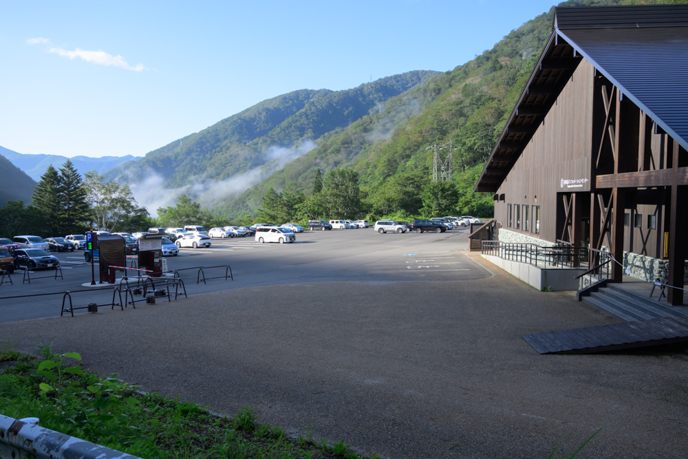 谷川岳インフォメーションセンターの駐車場。多くの車が停まっており、朝の山々に霧がかかっている