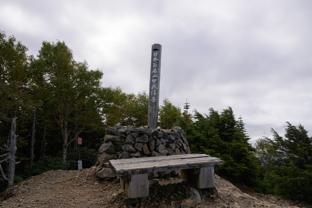「日本百名山 甲武信岳」の標柱と、石積みの前の木のベンチ