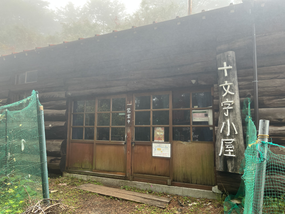 霧の中に建つ「十文字小屋」の看板がある木造の小屋