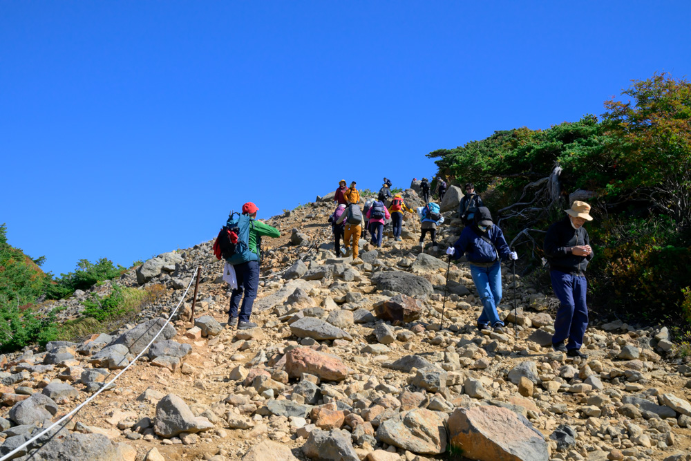 大きな石が転がる急な登山道を登る登山者たち。道の脇にロープが張られている