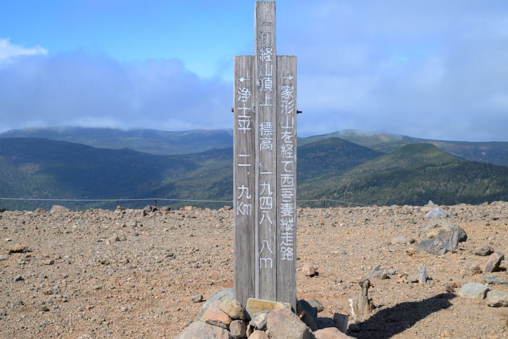 「一切経山頂上 標高1948.8m」と書かれた木の標柱。奥に山並みが広がる