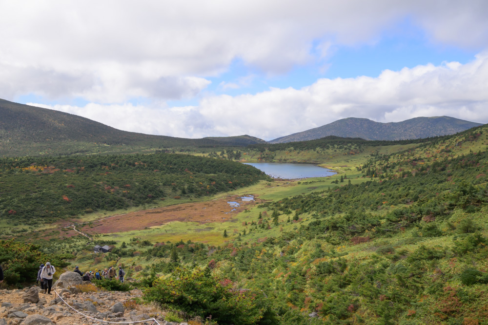 石の多い登山道を歩く登山者と、奥に見える沼と草紅葉の湿原