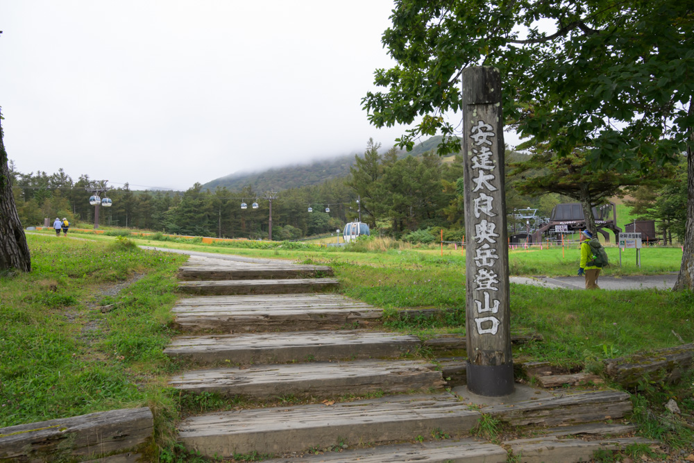 「安達太良奥岳登山口」と書かれた木の標柱と木の階段。奥にゴンドラと雲のかかった山が見える