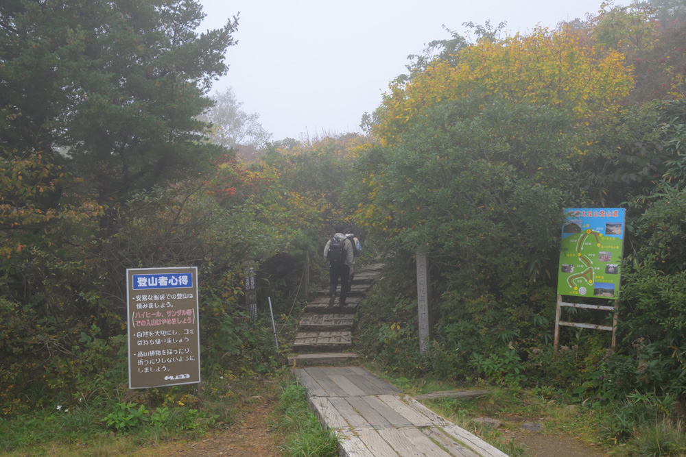 霧の中、木道と木の階段から始まる登山道の入口。「登山者心得」の看板が立っている