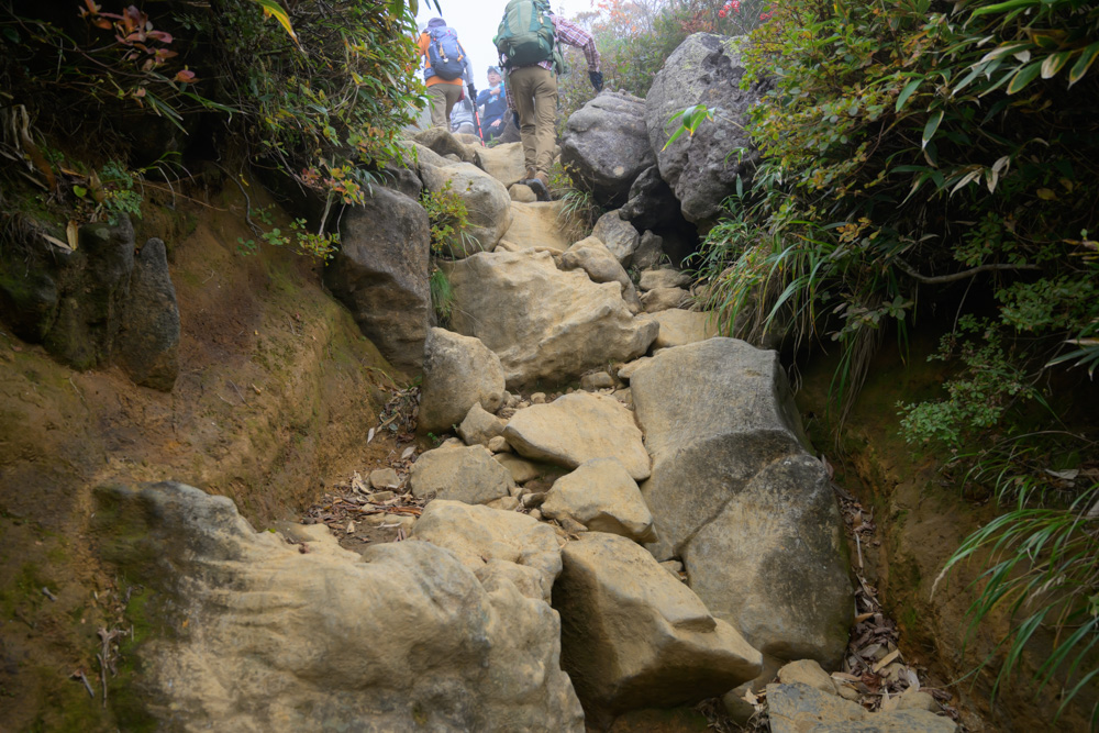 大きな岩が重なる登山道を登る登山者