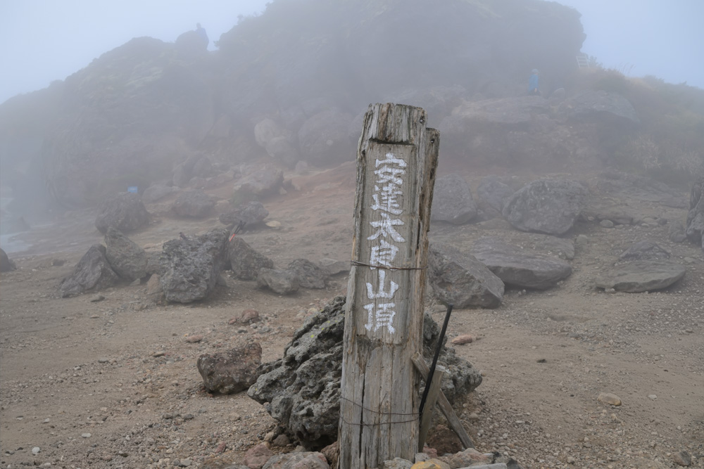 霧の中に立つ「安達太良山頂」と書かれた木の標柱。奥に岩場が見える