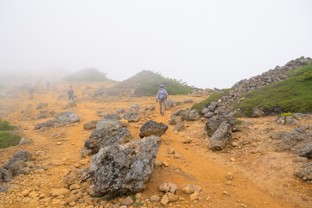 霧に包まれた、黄土色の砂礫と岩が広がる道を歩く登山者